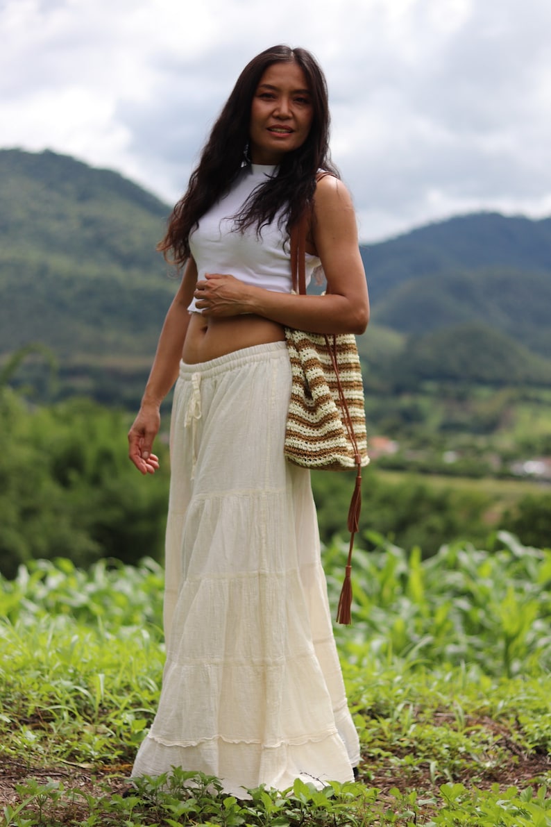 K&ouml;nnte beinhalten: Eine Frau tr&auml;gt ein wei&szlig;es Crop-Top und einen wei&szlig;en, mehrlagigen Maxirock mit einer braunen und wei&szlig;en geh&auml;kelten Tasche. Sie steht auf einem Feld mit gr&uuml;nem Gras und Bergen im Hintergrund.