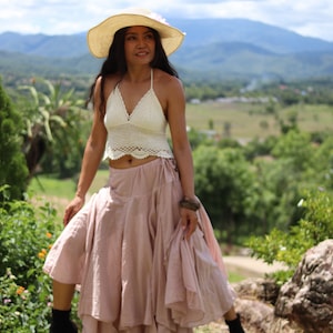 May include: A woman wearing a white crochet halter top and a long, flowy pink skirt. She is standing in a field with mountains in the background and is wearing a wide-brimmed straw hat.