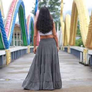 May include: A woman wearing a white crop top and a long, flowing gray skirt walks across a bridge decorated with colorful, serpentine sculptures.