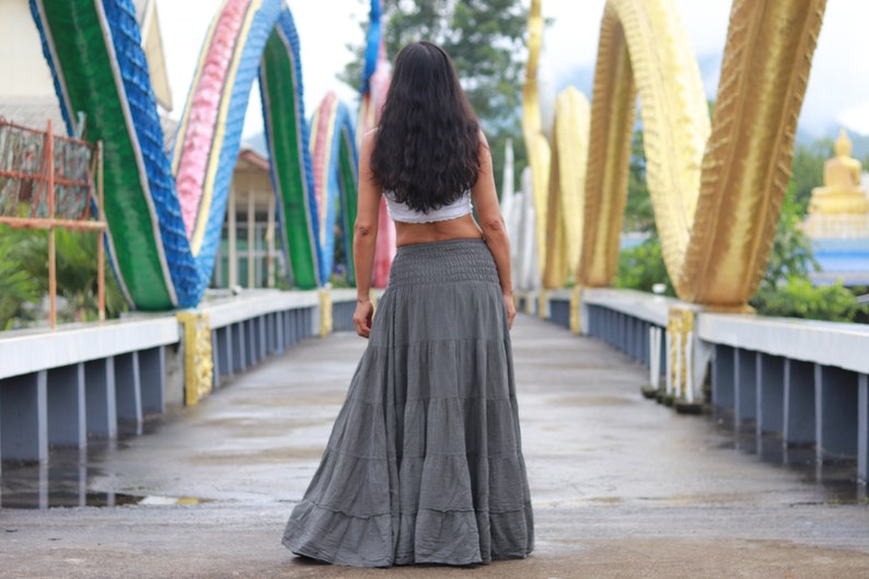 May include: A woman wearing a white crop top and a long, flowing gray skirt walks across a bridge decorated with colorful, serpentine sculptures.