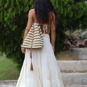 May include: A woman wearing a white tiered maxi skirt and a brown and white striped crocheted bag with a brown tassel.