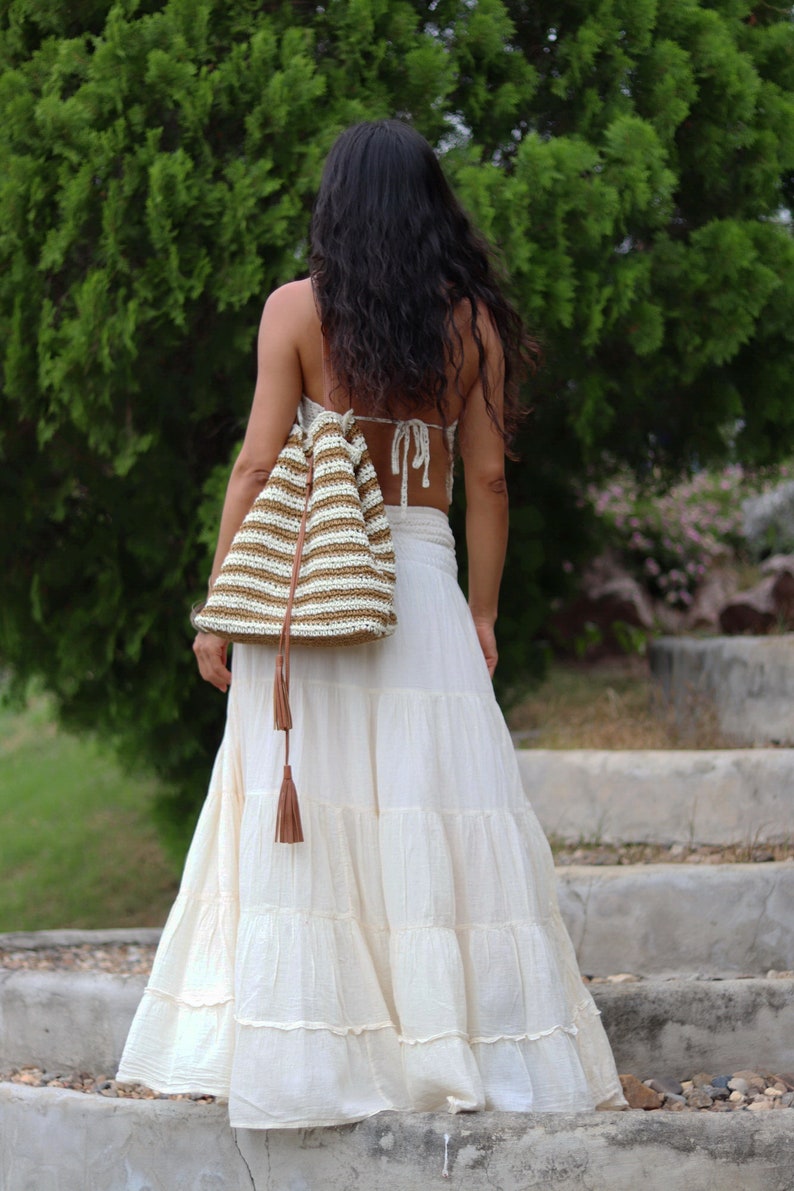 May include: A woman wearing a white tiered maxi skirt and a brown and white striped crocheted bag with a brown tassel.