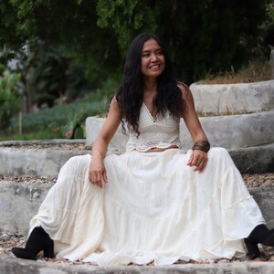 May include: A woman wearing a white crochet top and a long white skirt sits on a set of concrete steps. She is smiling and looking to the right.