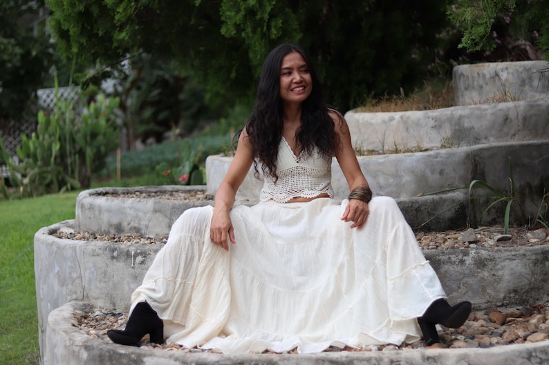 May include: A woman wearing a white crochet top and a long white skirt sits on a set of concrete steps. She is smiling and looking to the right.