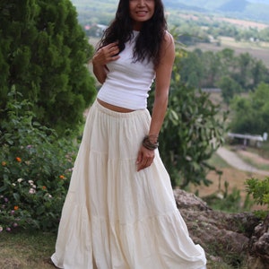May include: A woman wearing a white sleeveless top and a long, tiered, cream-coloured skirt. She is standing on a grassy hillside with a mountain range in the background.