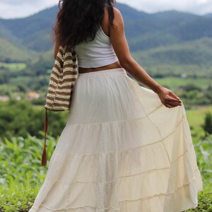 K&ouml;nnte beinhalten: Eine Frau tr&auml;gt ein wei&szlig;es Crop-Top und einen langen, wei&szlig;en, geteilten Rock. Sie h&auml;lt eine braune und wei&szlig;e geh&auml;kelte Tasche mit einem langen Quasten. Sie steht auf einem Feld mit gr&uuml;nem Gras und Bergen im Hintergrund.