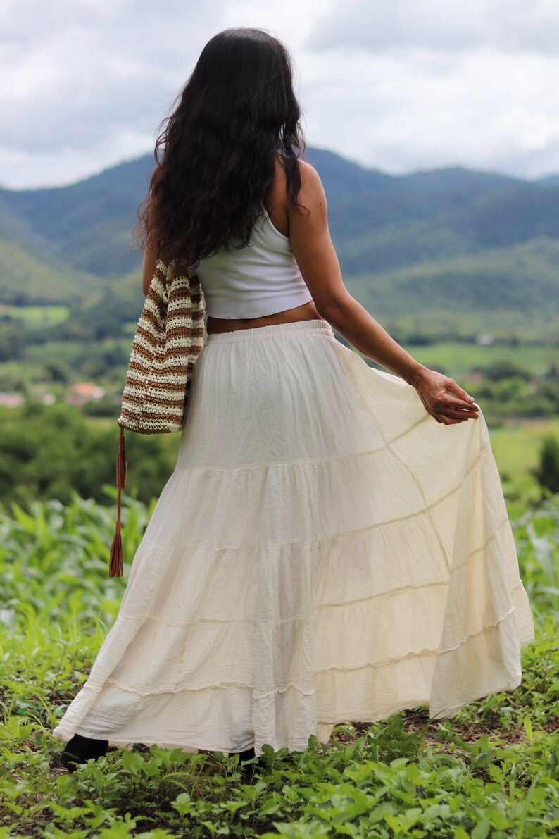 K&ouml;nnte beinhalten: Eine Frau tr&auml;gt ein wei&szlig;es Crop-Top und einen langen, wei&szlig;en, geteilten Rock. Sie h&auml;lt eine braune und wei&szlig;e geh&auml;kelte Tasche mit einem langen Quasten. Sie steht auf einem Feld mit gr&uuml;nem Gras und Bergen im Hintergrund.