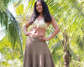 May include: A woman wearing a white crochet top and a brown tiered maxi skirt stands in front of a lush green palm tree grove.