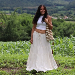 K&ouml;nnte beinhalten: Eine Frau tr&auml;gt ein wei&szlig;es Crop-Top und einen wei&szlig;en, mehrlagigen Maxirock mit einer braunen und wei&szlig;en geh&auml;kelten Tasche.