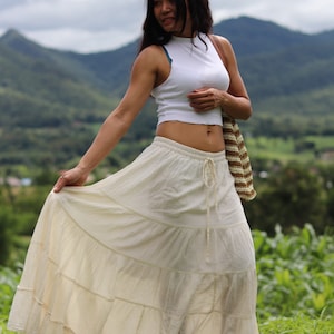 K&ouml;nnte beinhalten: Eine Frau tr&auml;gt ein wei&szlig;es Crop-Top und einen langen, wei&szlig;en, geteilten Rock. Sie steht auf einem Feld mit Bergen im Hintergrund.