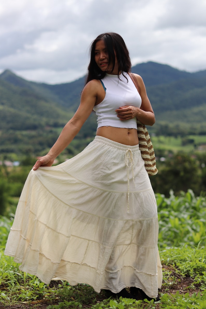K&ouml;nnte beinhalten: Eine Frau tr&auml;gt ein wei&szlig;es Crop-Top und einen langen, wei&szlig;en, geteilten Rock. Sie steht auf einem Feld mit Bergen im Hintergrund.