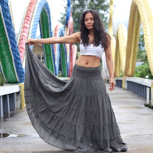 May include: A woman wearing a white crop top and a long, flowing gray skirt stands on a bridge. The bridge is decorated with colorful arches.