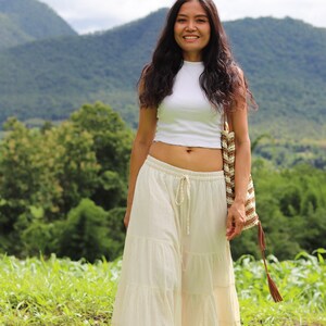 K&ouml;nnte beinhalten: Eine Frau tr&auml;gt ein wei&szlig;es Crop-Top und einen wei&szlig;en, mehrlagigen Maxirock. Sie h&auml;lt eine braune und wei&szlig;e geh&auml;kelte Tasche mit einem braunen Quasten. Sie steht auf einem Feld mit gr&uuml;nem Gras und Bergen im Hintergrund.