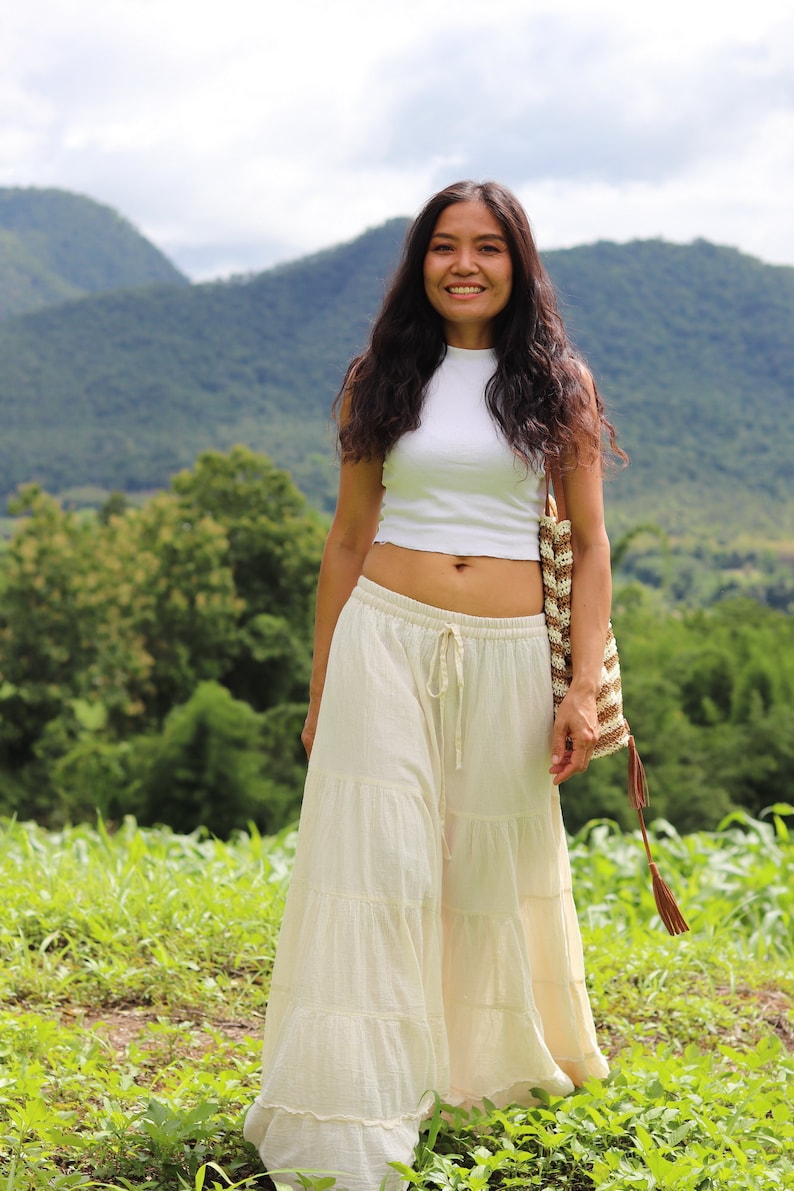 K&ouml;nnte beinhalten: Eine Frau tr&auml;gt ein wei&szlig;es Crop-Top und einen wei&szlig;en, mehrlagigen Maxirock. Sie h&auml;lt eine braune und wei&szlig;e geh&auml;kelte Tasche mit einem braunen Quasten. Sie steht auf einem Feld mit gr&uuml;nem Gras und Bergen im Hintergrund.