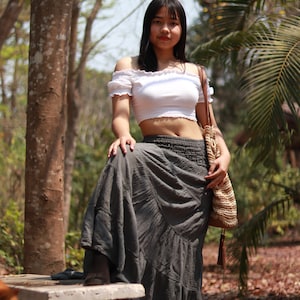 May include: A woman wearing a white off-the-shoulder crop top and a long gray skirt. She is standing in a forest setting with a brown woven bag slung over her shoulder.