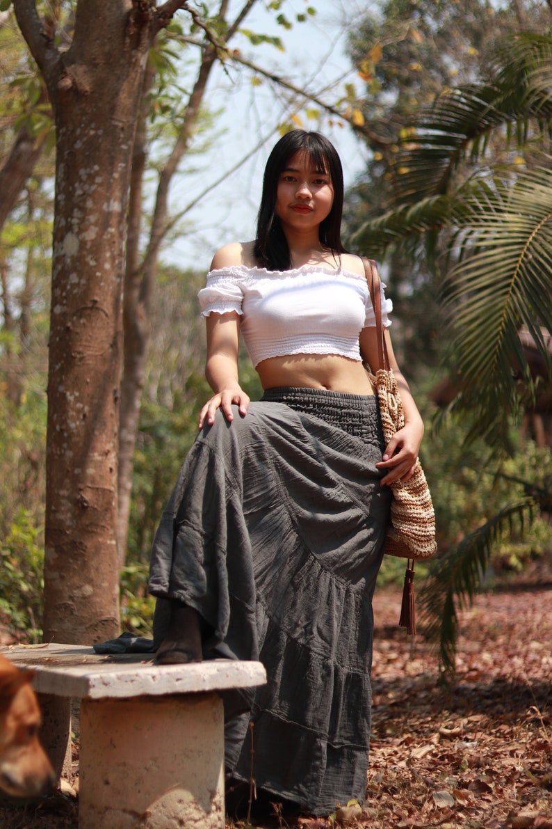 May include: A woman wearing a white off-the-shoulder crop top and a long gray skirt. She is standing in a forest setting with a brown woven bag slung over her shoulder.