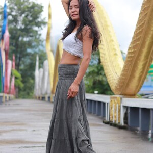 May include: A woman wearing a white crop top and a gray tiered maxi skirt. She is standing on a bridge with a golden structure in the background.