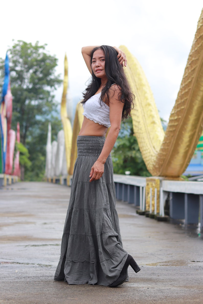 May include: A woman wearing a white crop top and a gray tiered maxi skirt. She is standing on a bridge with a golden structure in the background.