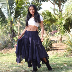 May include: A woman wearing a white crop top and a long, dark blue skirt with a ruffled hem. She is standing in a garden with lush green foliage.