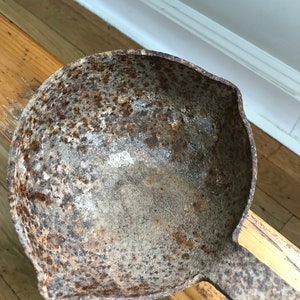 May include: A rusty metal ladle with a long handle. The ladle is made of cast iron and has a worn, aged appearance.