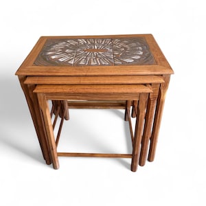 May include: Set of three nesting tables with a warm wood finish. The top table features a decorative tile inlay with a radial pattern in shades of brown, white, and gray. The tables are of varying sizes, designed to fit neatly together.