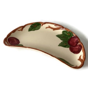 May include: A white ceramic serving dish shaped like a crescent moon with a brown twig design and red and green apples.
