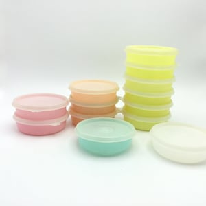 May include: A set of six round plastic containers with lids in various pastel colors, including pink, peach, light green, and yellow. The containers are stacked on top of each other.