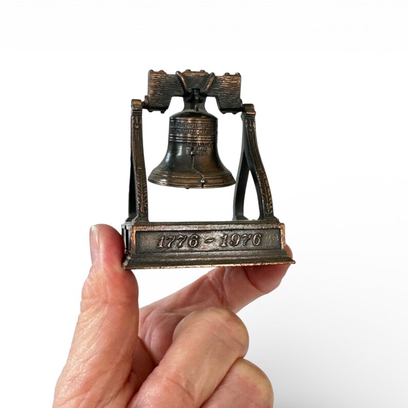 May include: A bronze-colored miniature Liberty Bell replica. The bell is suspended within a frame and sits on a base with the engraved text "1776-1976". The bell is held in a hand against a white background.