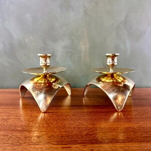 May include: Two gold-colored candle holders. Each has a round base with three curved legs and a small, flat disc to catch wax. The candle holders are made of a shiny metal and are displayed on a wooden surface.