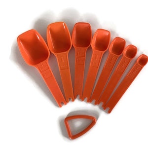 May include: A set of seven orange measuring spoons, arranged in a fan shape, with a small orange ring. Each spoon is labeled with a measurement, including 1 tbsp, 1 tsp, and fractions thereof. The spoons are made of plastic.