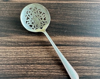 May include: A silver-toned serving spoon featuring a circular bowl with an intricate, cut-out filigree pattern and a slender, elongated handle with subtle linear detailing along the edges, designed for serving food items.