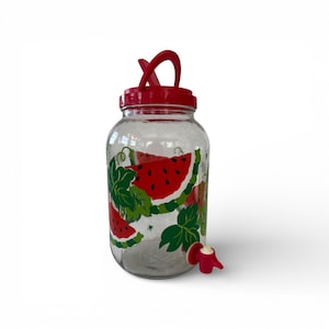 May include: A clear glass beverage dispenser with a red lid and spigot. The dispenser is decorated with watermelon slices and green leaves. The lid has a red handle. The spigot is red and white.
