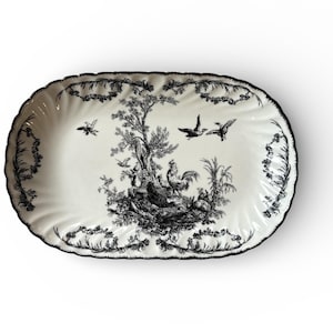 May include: An oval serving platter with a black and white toile design. The platter features a detailed pastoral scene with birds, trees, and farm animals. The rim is scalloped and trimmed with a black floral pattern. A decorative piece for serving.