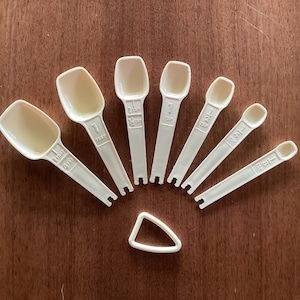 May include: A set of seven white plastic measuring spoons with black markings. The spoons are arranged in a fan shape, with the largest spoon at the top and the smallest spoon at the bottom. The spoons are labeled with the following measurements: 1 tsp, 1/2 tsp, 1/4 tsp, 1/8 tsp, 1/16 tsp, 1/32 tsp, and 1/64 tsp.