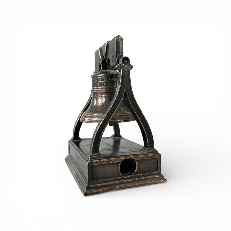 May include: A bronze-colored pencil sharpener shaped like the Liberty Bell. The bell is suspended within a frame, resting on a rectangular base with a pencil-sized hole. The words "Liberty Bell" are visible on the bell.