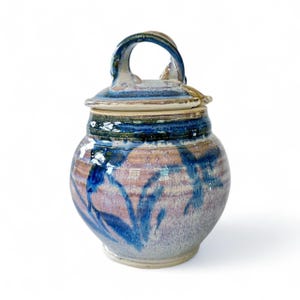 May include: Handmade ceramic jar with lid. The jar features a bulbous shape with a handle on top. The exterior is painted with blue and purple abstract designs. The lid has a matching blue and purple color scheme.