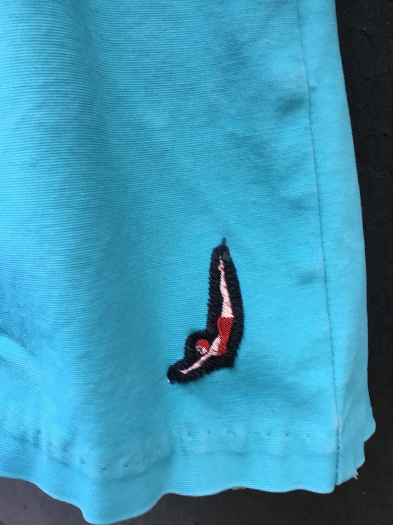 May include: A turquoise blue pair of shorts with a red, white, and black embroidered image of a diver on the right leg.