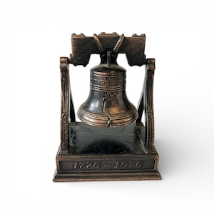 May include: A bronze-colored Liberty Bell replica with a dark base and wooden-textured yoke. The bell features engraved text and is supported by ornate columns. The base displays the numbers "1776 - 1976".
