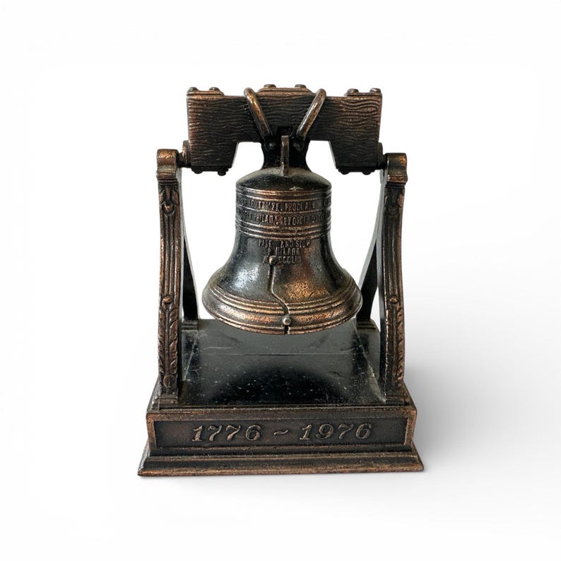 May include: A bronze-colored Liberty Bell replica with a dark base and wooden-textured yoke. The bell features engraved text and is supported by ornate columns. The base displays the numbers "1776 - 1976".