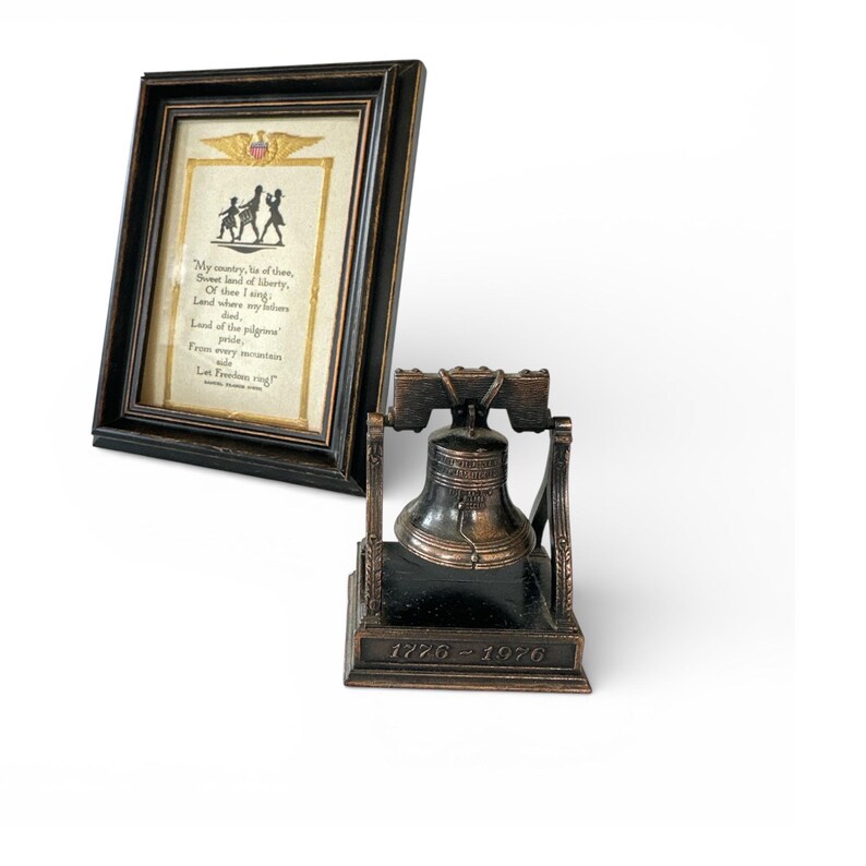 May include: A framed print with a patriotic theme, featuring a poem and silhouette figures. Beside it, a bronze-colored Liberty Bell replica with the date 1776-1976. The frame is dark brown, and the bell sits on a black base.