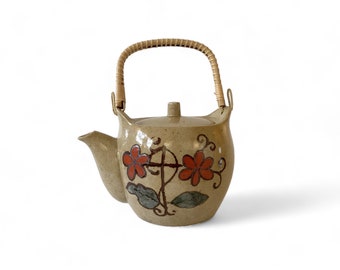 May include: A beige ceramic teapot with a brown woven handle. The teapot has a floral design with red flowers and green leaves.