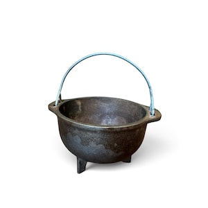 May include: A dark gray cast iron cauldron with a rounded body, three short legs, and two side handles. A thin, arched metal bail handle is attached to the top, extending upwards. The surface of the pot has a textured, matte finish.