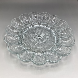 May include: Clear glass deviled egg serving platter. The round platter has a scalloped edge with individual egg compartments. The center of the platter has a textured, dotted design. A small hole is in the center of the platter.