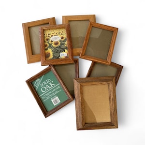 May include: A collection of rectangular wooden photo frames in various shades of brown. One frame contains a picture of sunflowers. One frame has a green label that reads "SOLID OAK 5x7 PHOTO FRAME". The frames are arranged on a white surface.