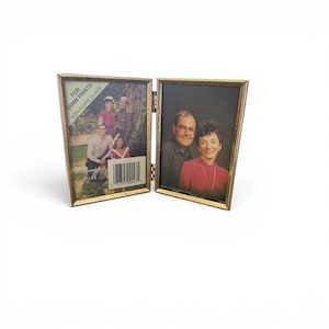 May include: A hinged, gold-colored metal photo frame, open to display two photographs. The frame is designed to hold 3.5 x 5 inch prints. One side features a family portrait, the other a couple. Text on the frame reads "FOR 35MM PRINTS NON-GLARE GLASS".