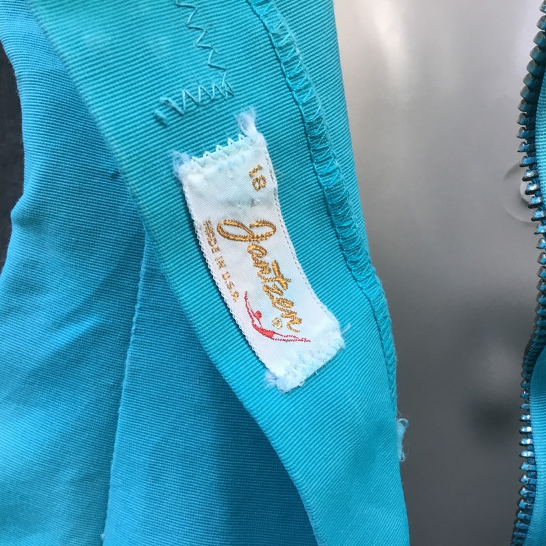 May include: A turquoise blue fabric with a white label that says "Jantzen" and "Size 18 Made in U.S.A.". The label has a red and white illustration of a woman diving.