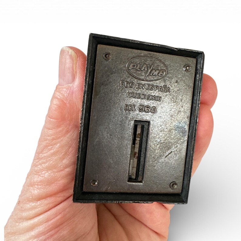 May include: A vintage metal pencil sharpener with the text "PLAY ME" and "FDO EN ESPA&Ntilde;A MADE IN SPAIN REF. 968". The sharpener has a rectangular shape with a dark frame and a silver-colored metal surface.