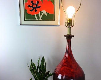 May include: A red and brown ceramic table lamp with a brass base and a white lampshade. The lamp is on a wooden table next to a potted plant.