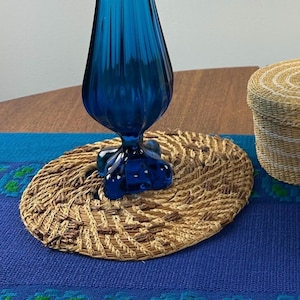 May include: A cobalt blue glass vase sits on a woven, oval-shaped placemat. The placemat is made of natural fibers and has a textured appearance. A small, woven basket is partially visible in the background. The scene is set on a blue tablecloth.