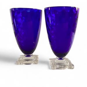 May include: Two cobalt blue drinking glasses with textured surfaces. The glasses have a flared shape and sit on clear, square-shaped bases. The glassware is set against a white background.